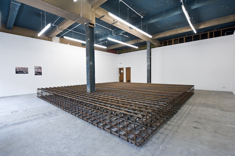 The view of a white walled. gallery space, concrete floor and exposed ceiling. Taking up a large area is a sculpture made from stacks of iron rods. On the wall behind are two black and white images.