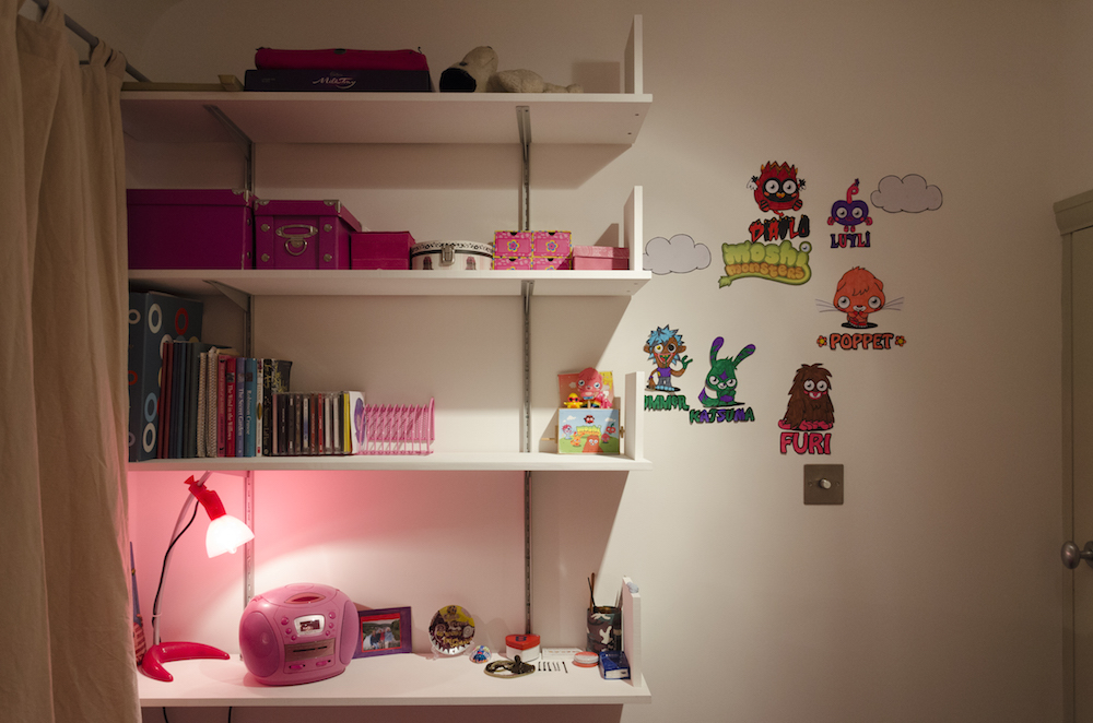 A photograph of some shelves in a bedroom. On the shelves are personal objects, likely belonging to a young girl. Many of the objects are pink, including a lamp and radio. There are stickers on the wall to the right of the shelves.