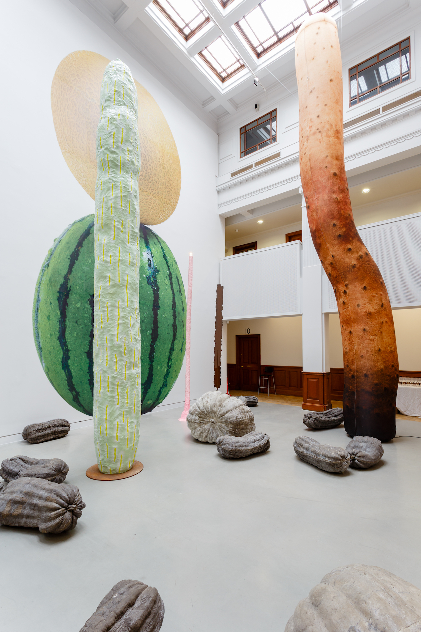 A series of sculptures arranged in the atrium of a gallery. Two huge cactus-like constructions rise vertically. On the floor are a selection of cast pumpkins and gourds in shades of grey.