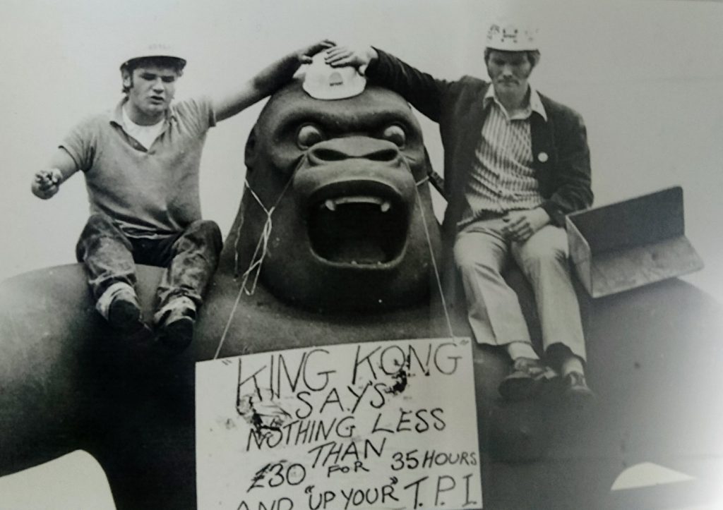 A black and white photograph of a fibreglass king kong sculpture, with a person sat on each shoulder. Around the gorilla's head hangs a sign about pay.