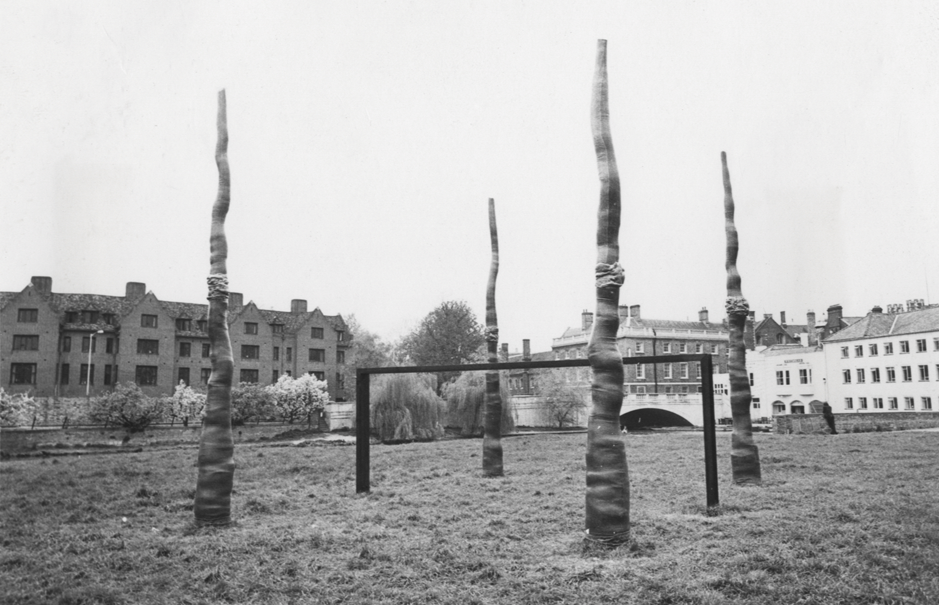 A black and white photograph of a public sculpture in a grassy area, with buildings at the edge of the image. The sculpture consists of four wiggly shapes reaching up to the sky, and in the centre a piece of metal that looks like football goalposts in black. 
