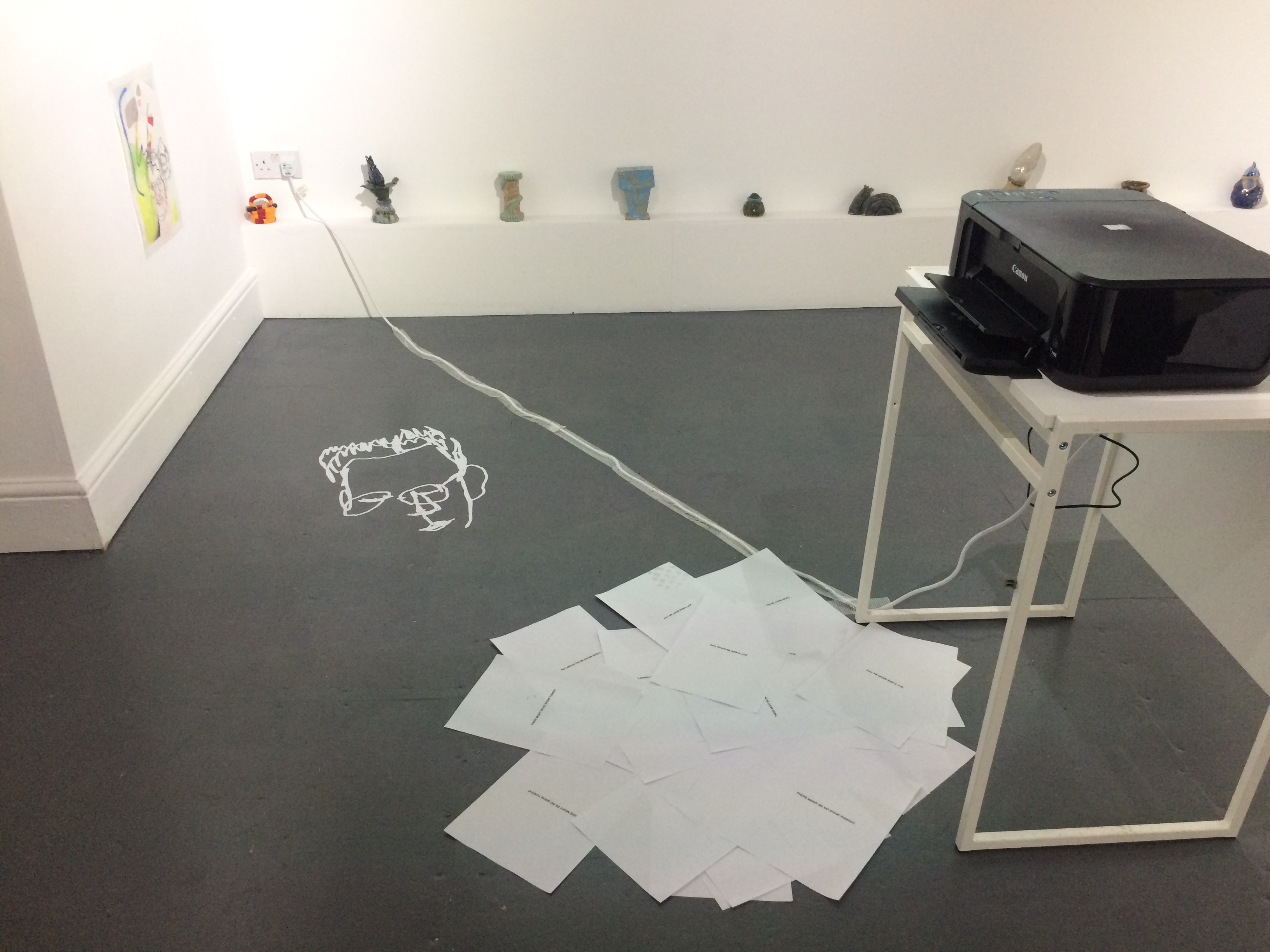 A gallery space with white walls and grey floor. A printer on a plinth prints out sheets of paper with small sentences of text on. There is a small chalk drawing on the floor. Around the edge of the room, small ceramics rest on a ledge.