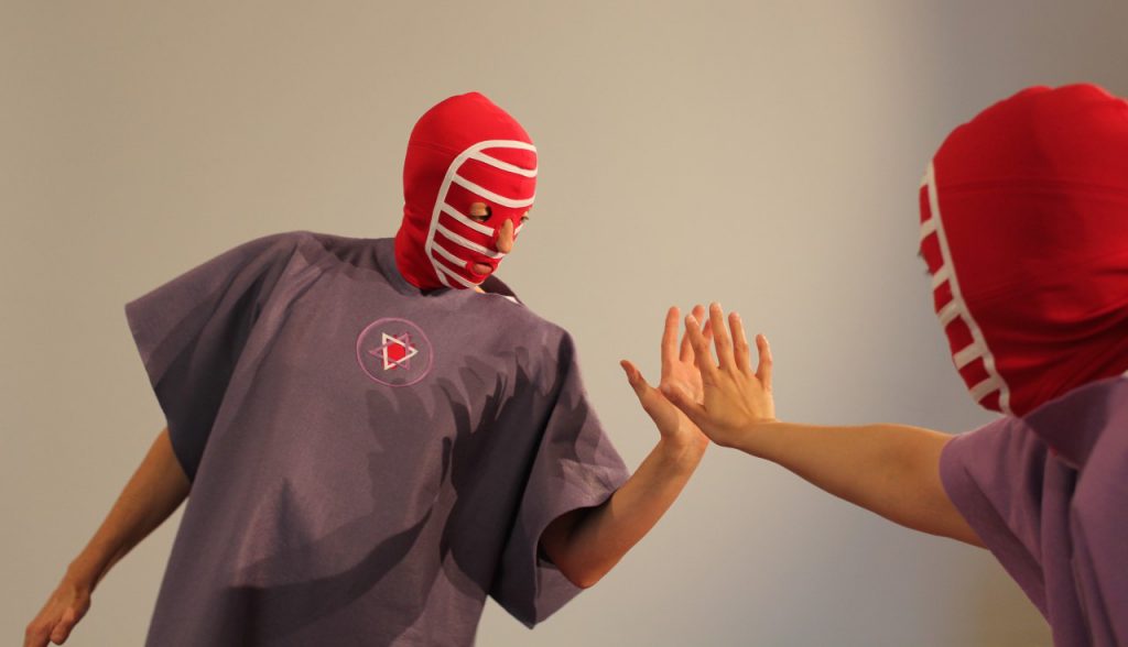 Two people dressed in red face masks and purple smocks face each other and reach our their hands which are almost touching.