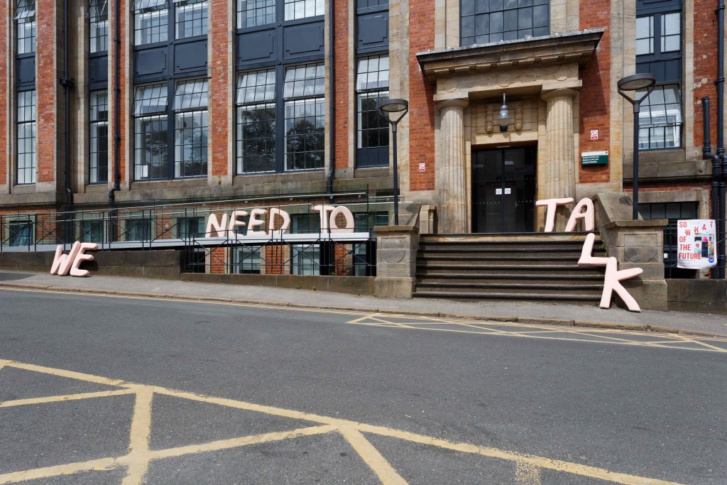A view of the exterior of a university building. A sculptural work spells out: 'we need to talk' across the front and steps.