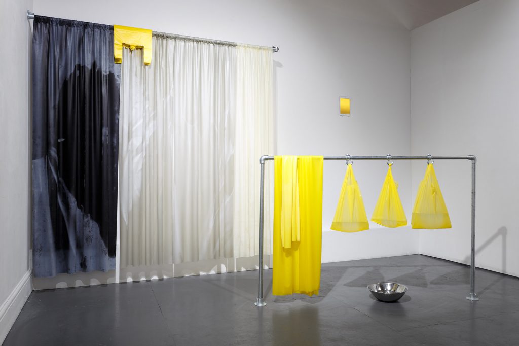 A view of a gallery with several sculptures forming an installation. The works resemble curtains and other household objects in clear plastic, grey and yellow.