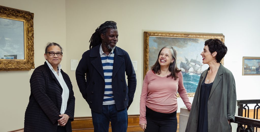 A group of four people stand in a gallery together, they are laughing and talking together.