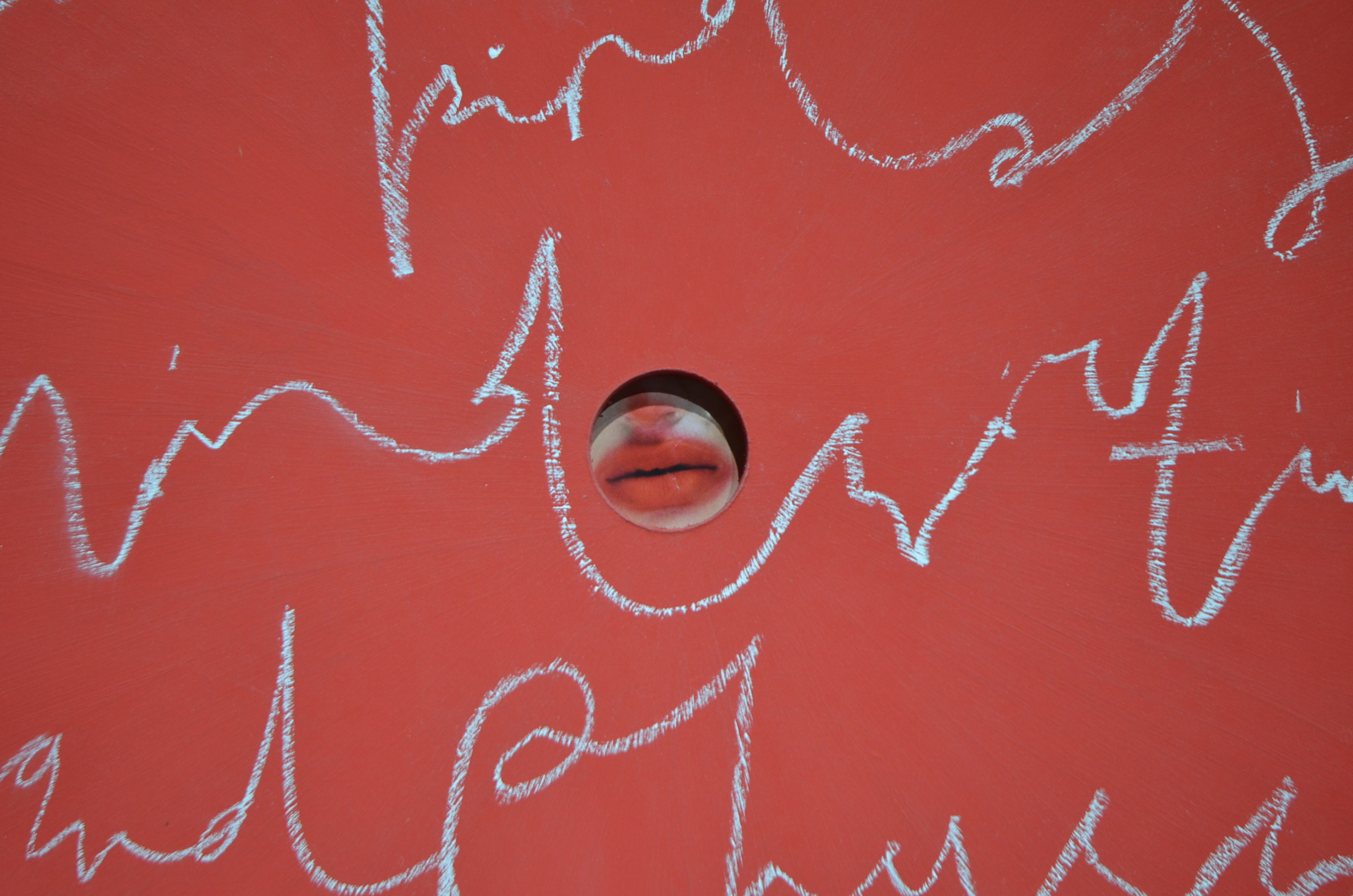 A red screen with writing in chalk. A hole in the board reveals a mouth, with the lips painted in the same shade of red.