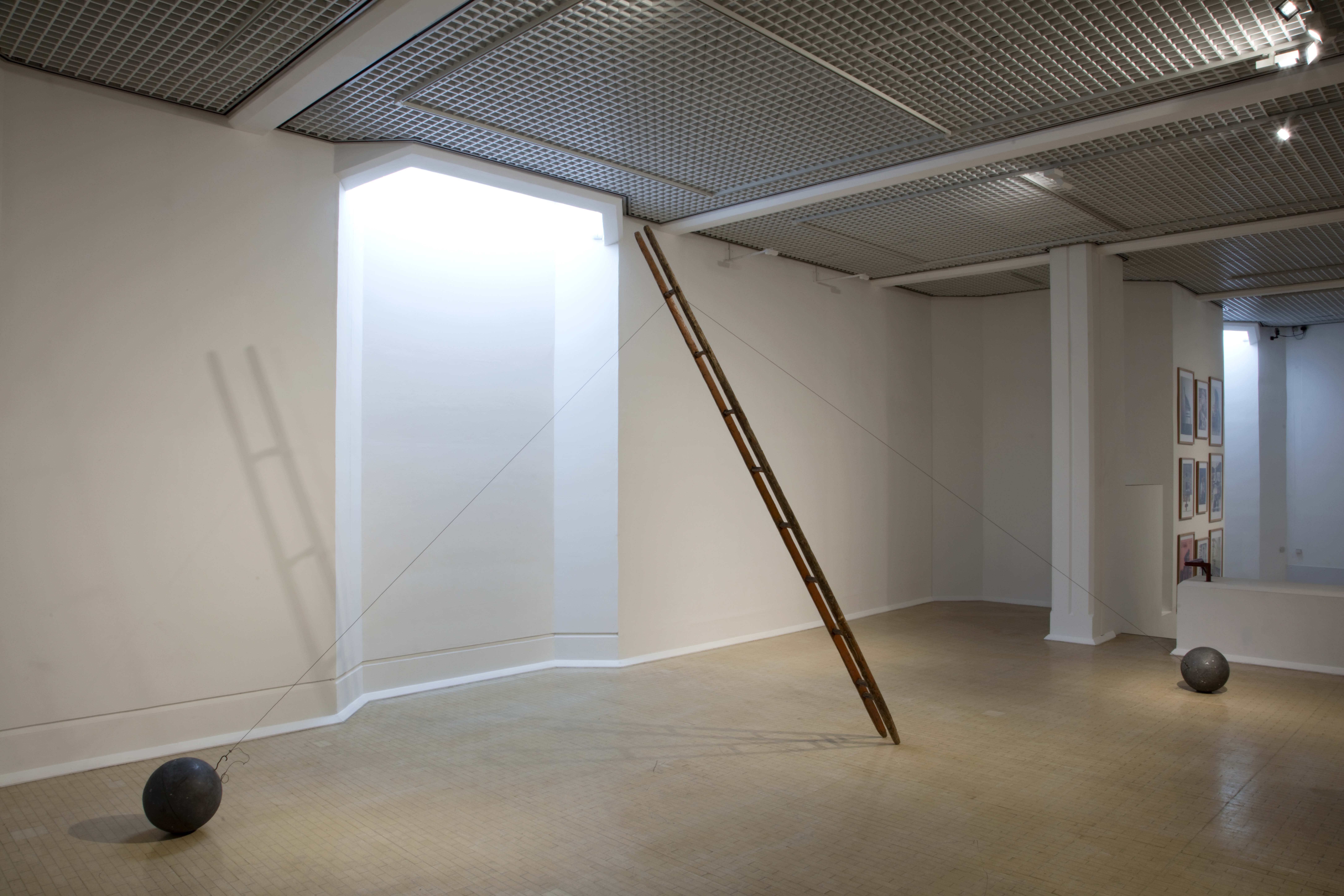 A gallery with white walls and wooden floor. Several sculptures can be seen in the room, the main focus is a ladder leading up to the ceiling. The ladder is pinned down by round ball weights and wire.