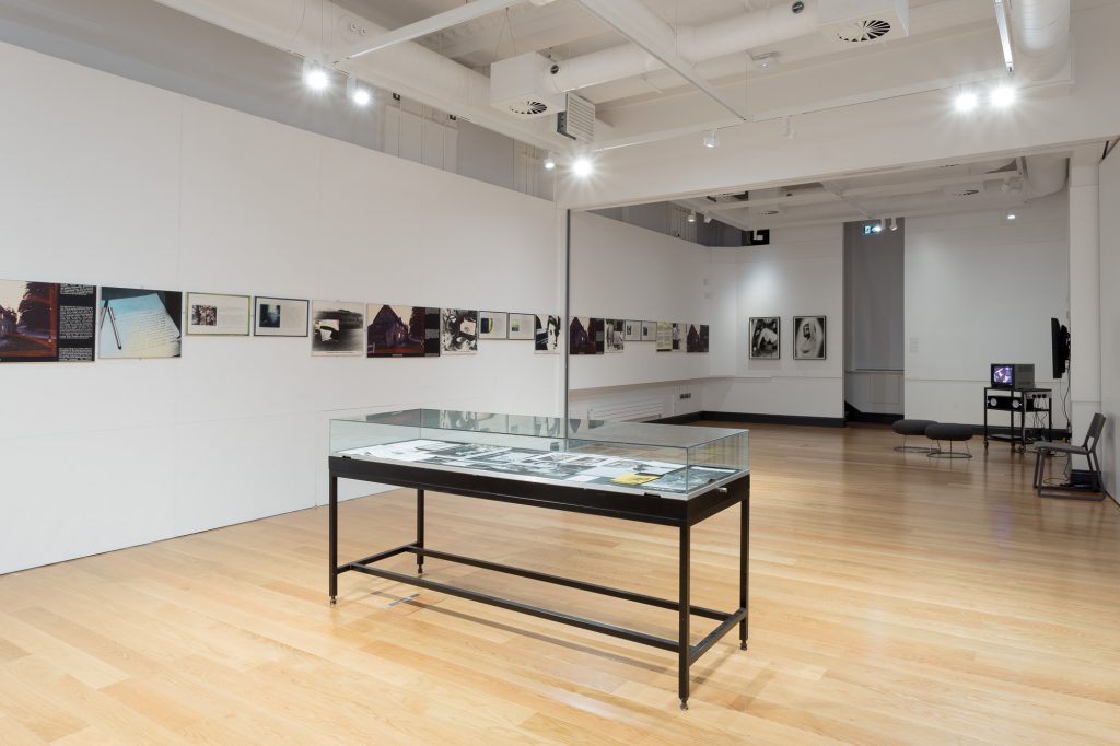 View of a gallery space with white walls and pale wooden floor. There are several works visible: a vitrine with black legs, photographic works mounted along the wall, and several small TVs screening artists' films.