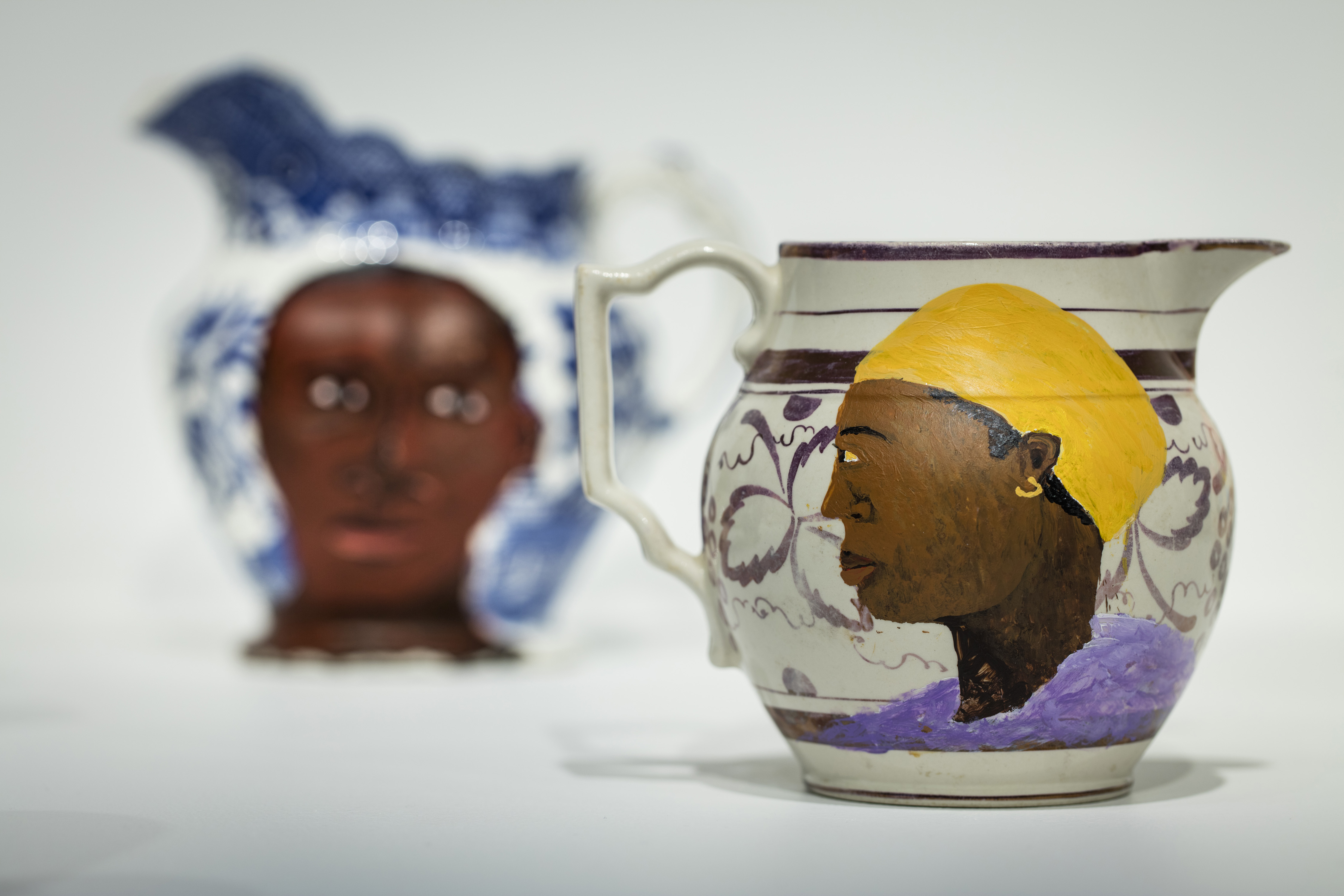 An image of two porcelain jugs. Both have been painted on by the artist, who has added tender portaits of black people to both. The paintings are vivid and overlaid on the existing patterns of the pieces.