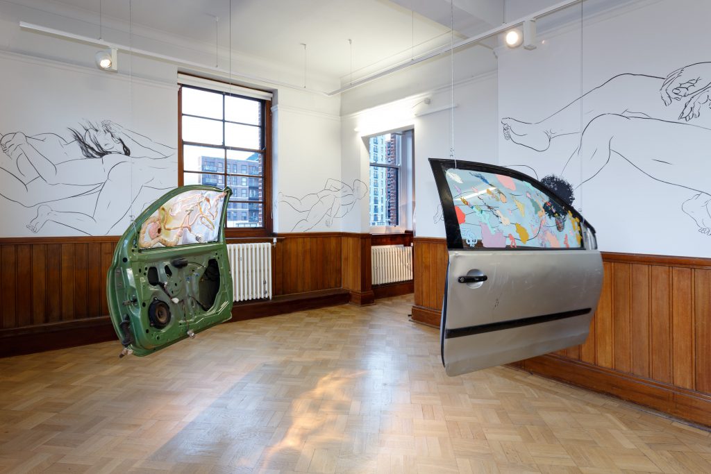 Two car doors hang in a gallery space. The glass windows of both have been painted with pastel shades and patterns. On the white walls in the background are drawings of sleeping and resting bodies and limbs.