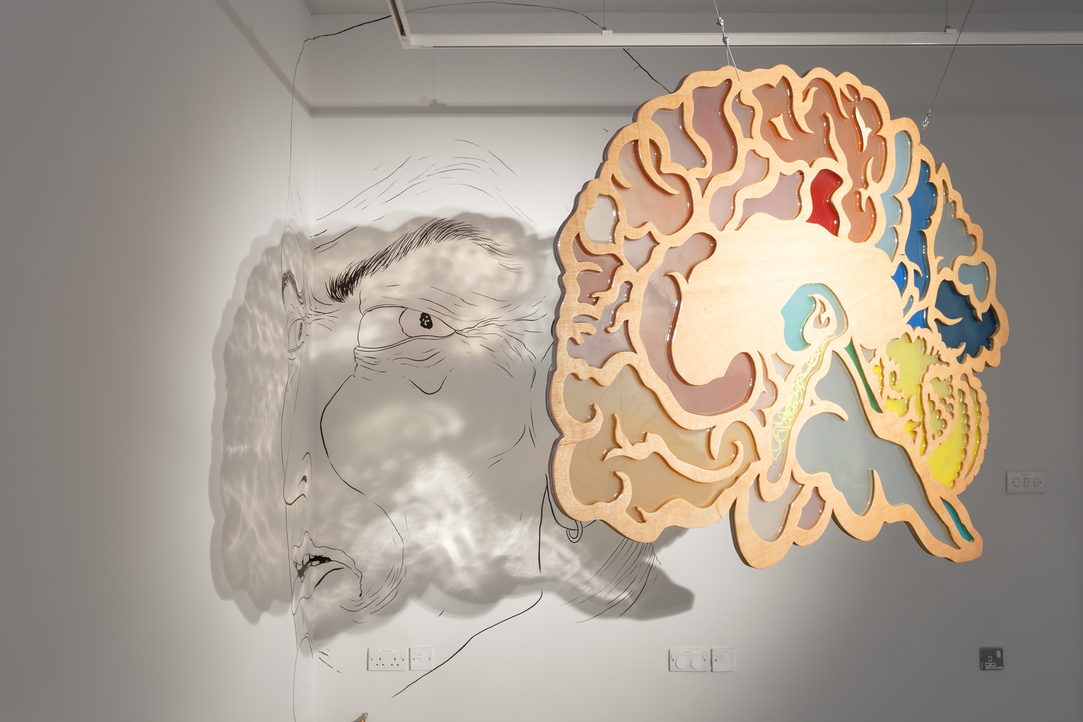 A hanging sculpture depicting a brain, with cloud like forms. it is made from plywood with coloured glass inserts. The shadow of the work falls on the wall behind, where it intertwines with a drawing in black on the white surface.