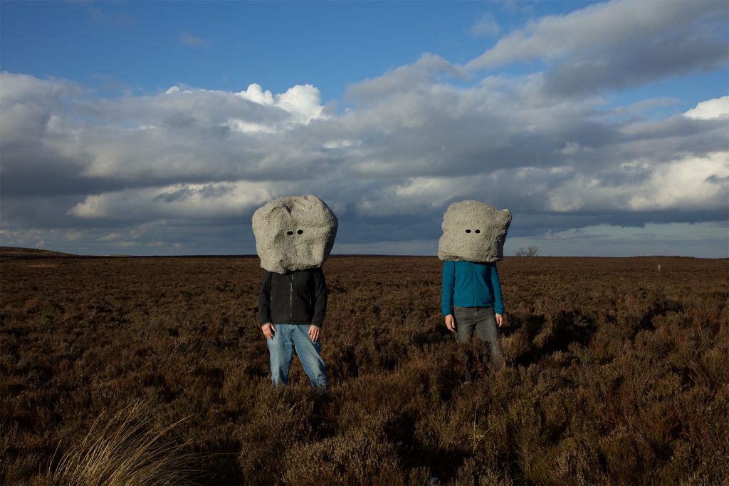 Two figures wearing large rock masks stand in a landscape.
