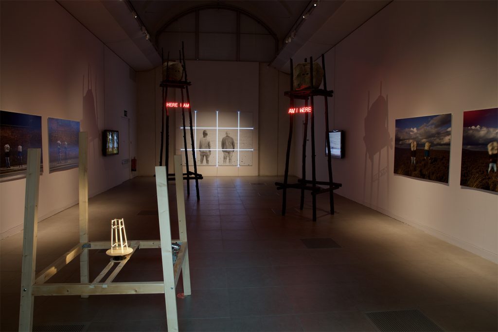 A darkened gallery containing a number of wall-based works and sculptures. Most visible are two tall platforms with rocks resting at the top. In neon are the words 'here I am' and 'am I here'.