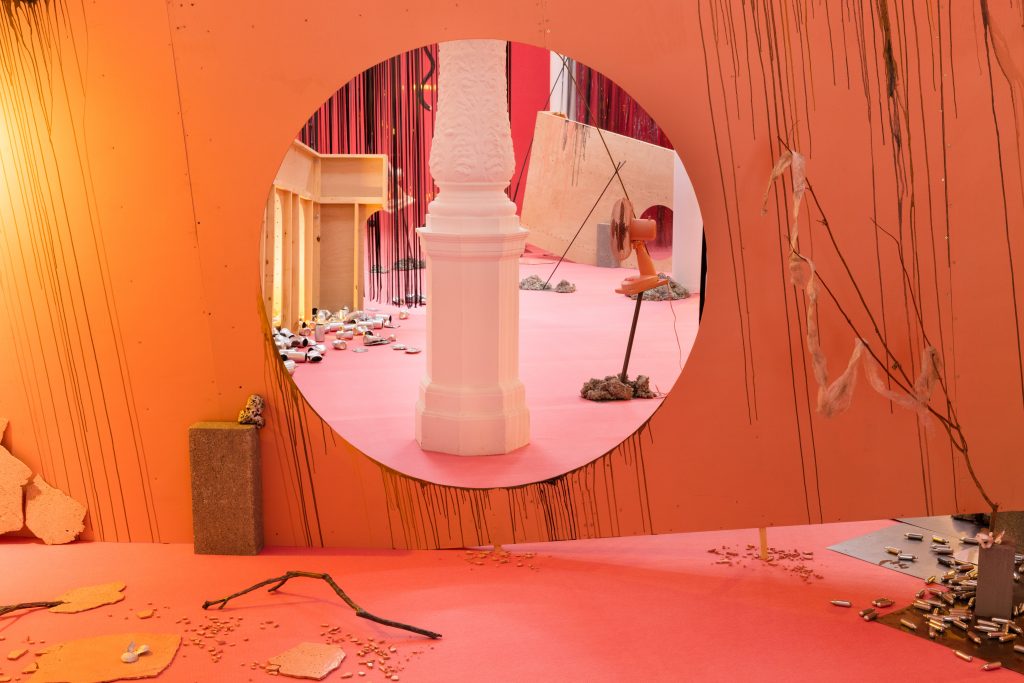 A room suffused with orange and peach light. In a circle, a view of a pillar is visible. Several objects such as a twig are visible resting on the floor.