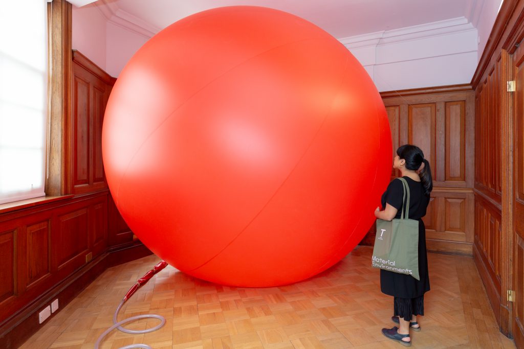 A room in a gallery with wooden floor and panelled walls. The room seems to be entirely filled by a large red ball. A visitor stands beside it.