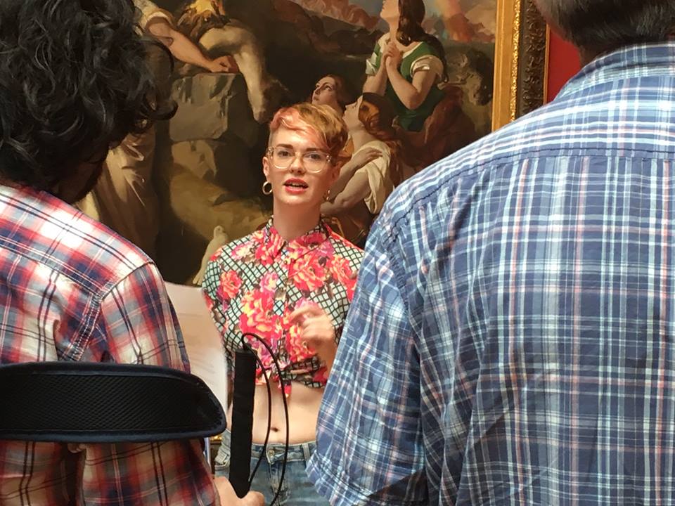 A white person with short pink hair and glasses stands in a gallery, they are reading a text to the audience.