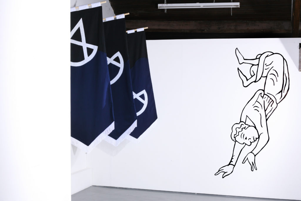 An installation with a series of flags in dark blue with a white design. On the white wall behind is an illustration of a person falling.