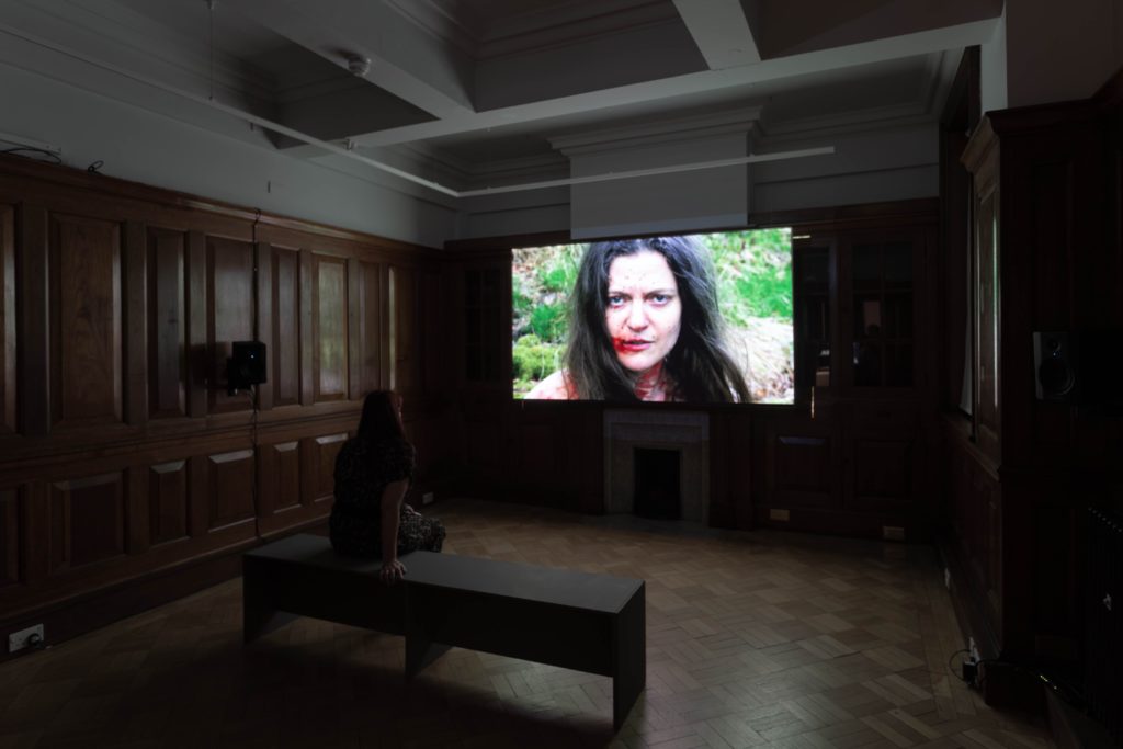 In a darkened room, a person watches an artist's film projected on the back wall. The image on screen shows a person with dark hair and blood round their mouth looking directly into the camera.