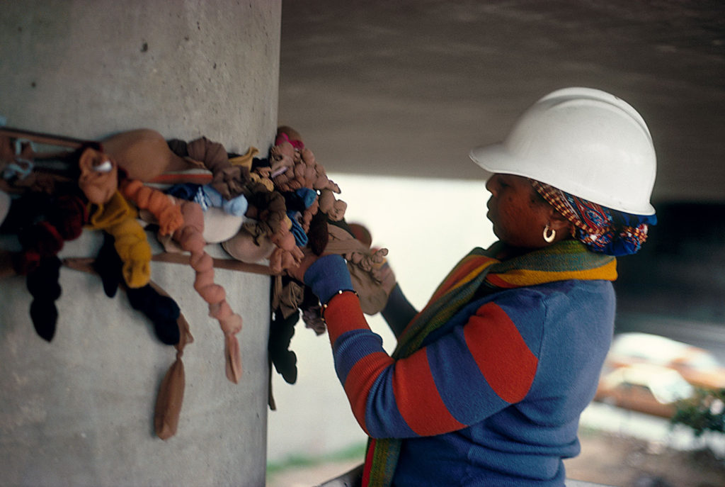 A black woman in a white hard hat and colourful jumper adjusts some sculptural fabric tied around a concrete column. The fabric looks like stuffed tights in different shades of brown, pink, black, yellow and blue.