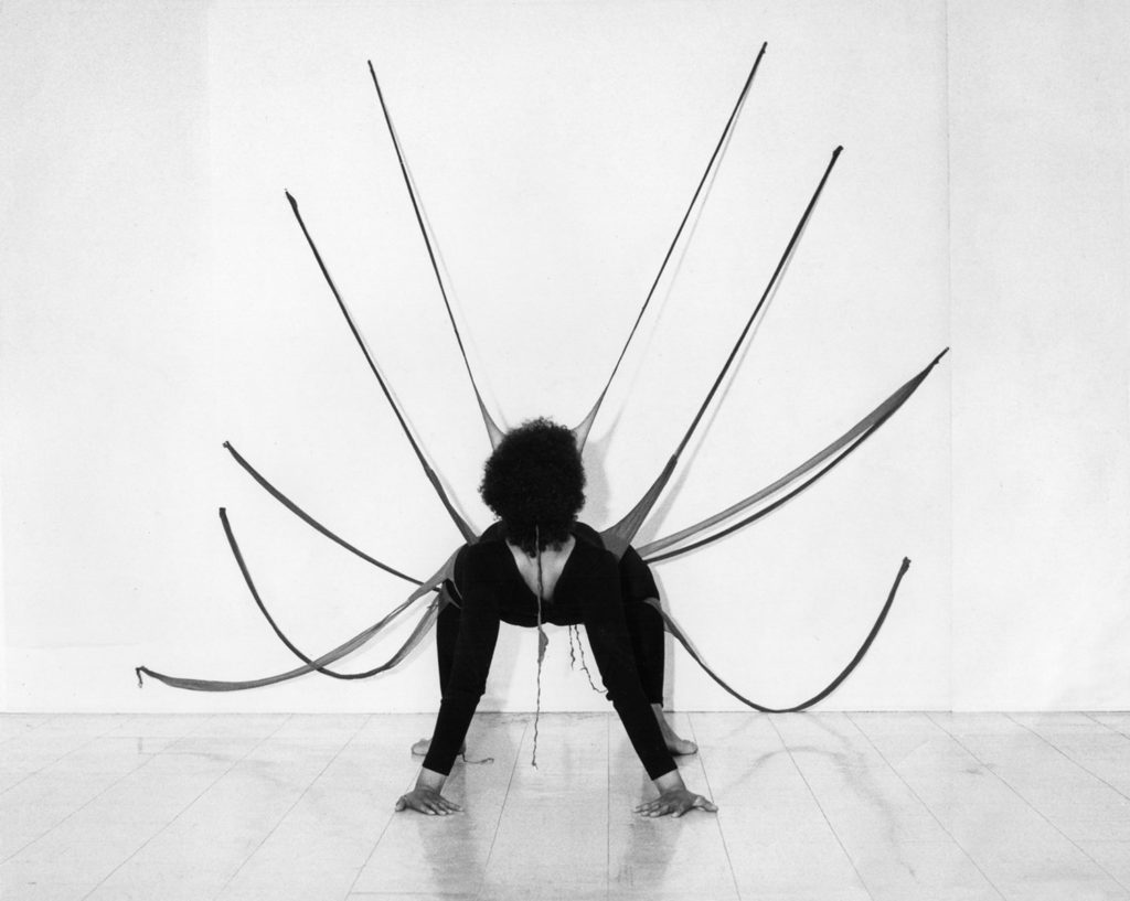 A black and white image of a body in the crab position. The back of their head and arms are visible to the viewer, while the rest of their body is foreshortened. Protruding from their body, and extending up onto the walls, are black fabric strings.