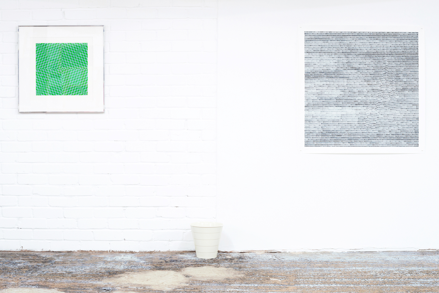 Two works hanging on a white gallery wall. They are both geometric in design, green on the left and grey on the right. Below them is a plastic white wastepaper bin.