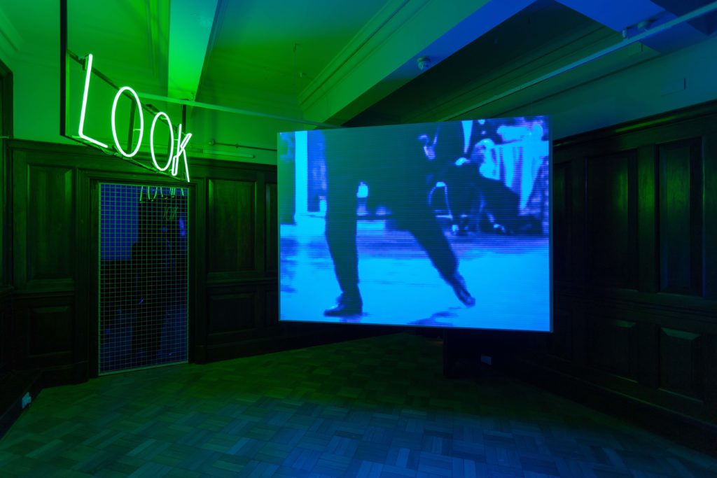 An artist's film screened in a gallery space. On screen is a view of a person's legs in blue light. In the surrounding gallery is a neon green work lighting the room.