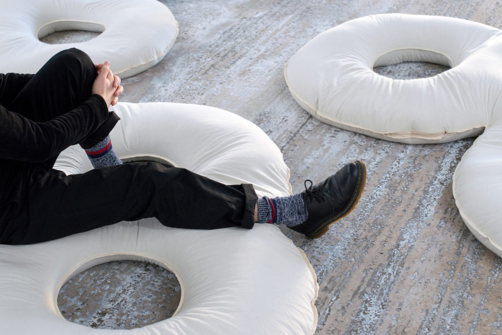 A person sitting on a donut shaped white cushion. Only their legs and feet are visible. They are dressed in black and grey.