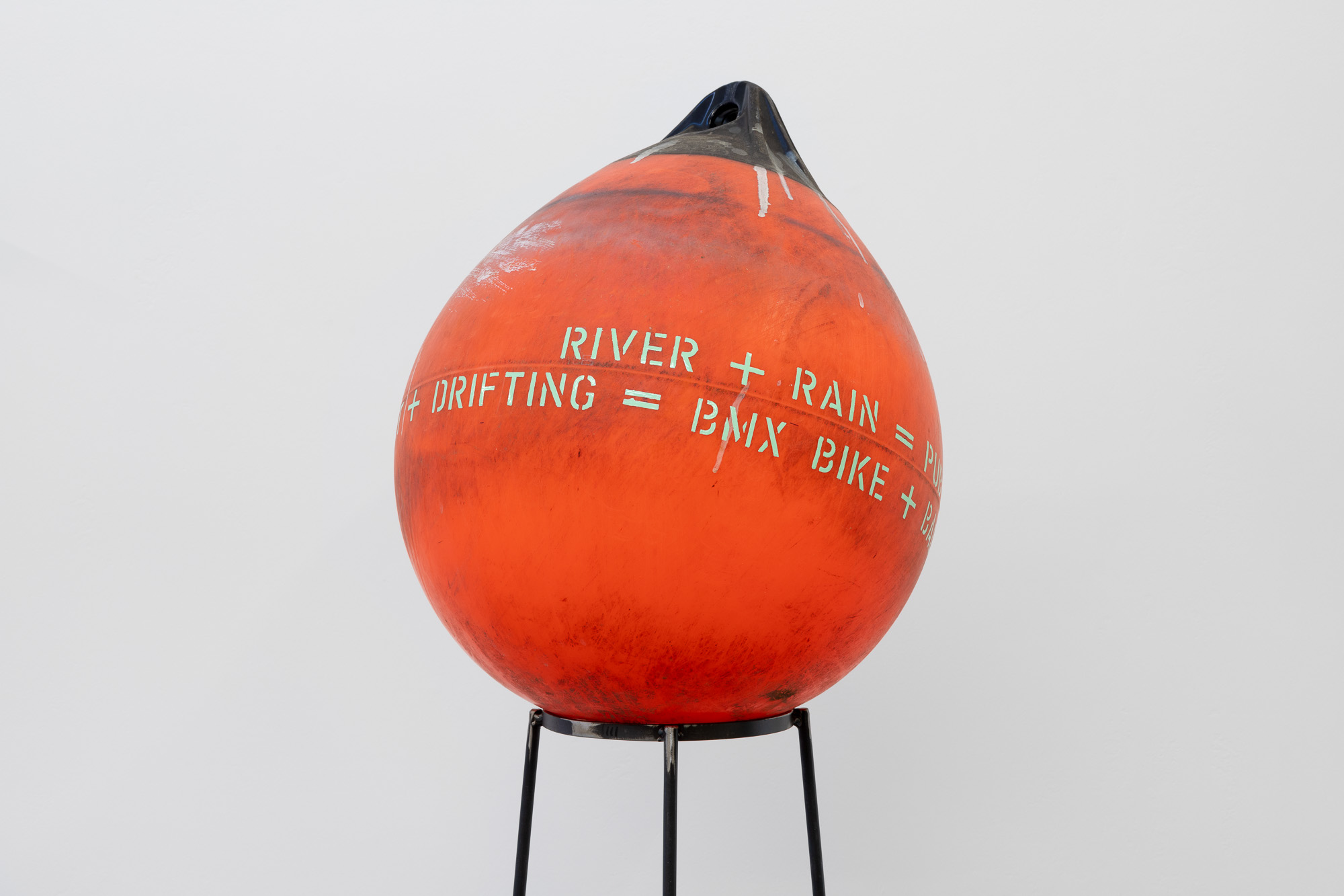 An orange buoy used at sea printed with the words 'RIVER + RAIN =' and on the second line '+ DRIFTING = BMX BIKE +'