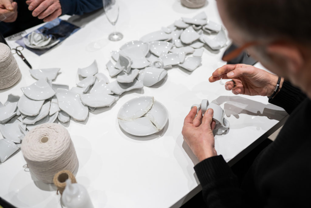 A table with pieces of white broken porcelain. People are sitting at the table, just their hands can be seen mending and piecing items back together in new formations.