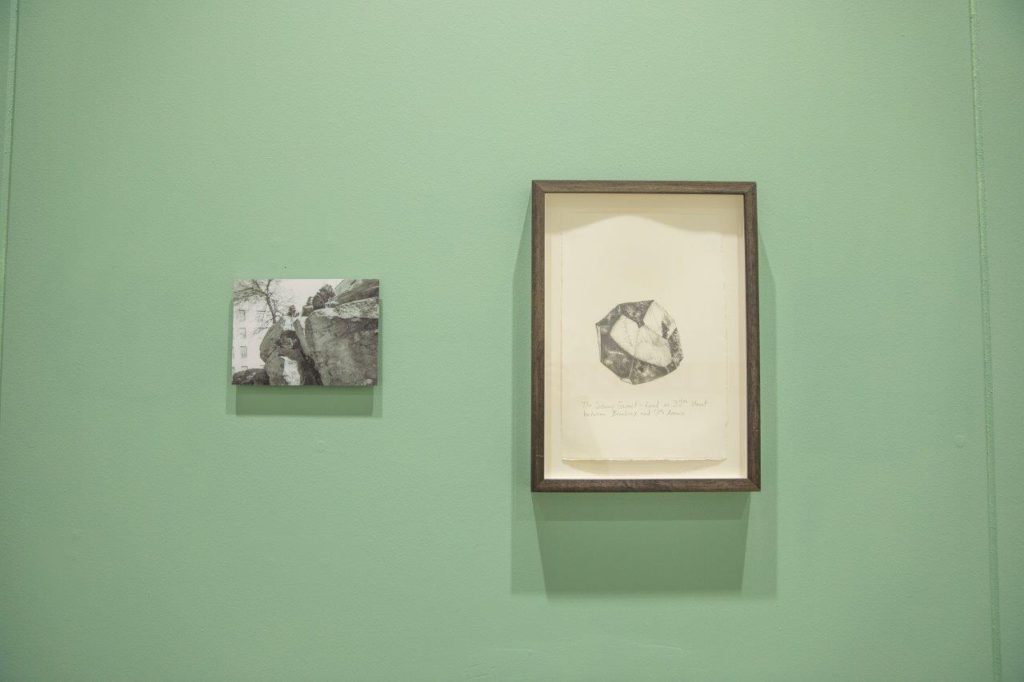 A green gallery wall featuring two artworks. On the left, a black and white image of rockface with a building in the background. On the right a drawing of a rock or mineral.