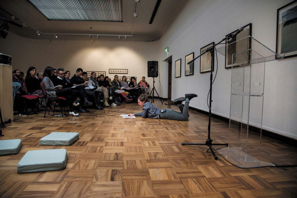 A gallery space with white walls and a wooden floor. Works can be seen hung on the walls. The seated audience look towards a person lay on the floor, reading, dressed in all grey.