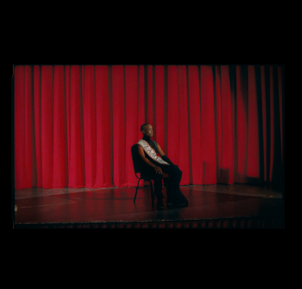A black person sits on a chair infront of a red theatre curtain. They are wearing a sash across their body.