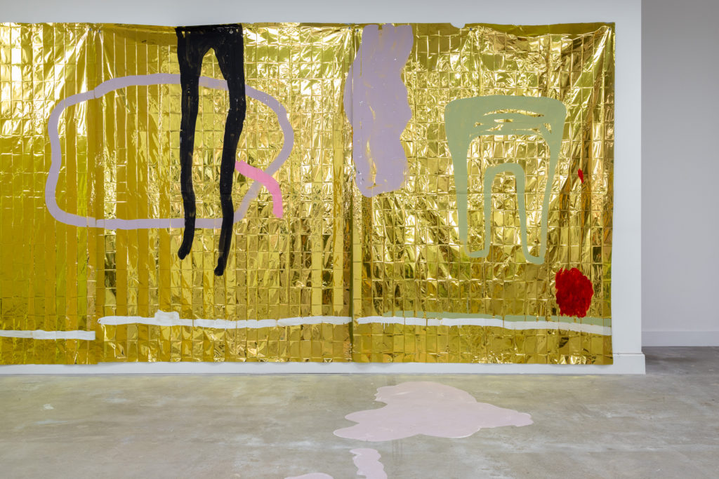 A view of an artwork in a gallery. It is a large wall hanging or mural, made from fabric. The background is shiny gold, and it features shapes that look like legs and bodies.