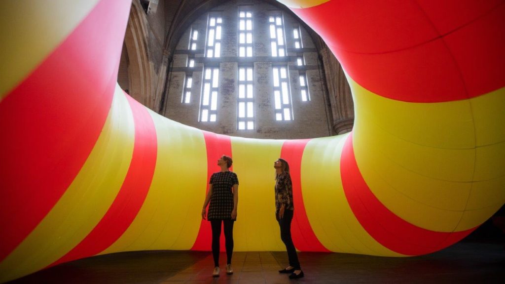 Two people stand inside a large inflatable artwork in shades of yellow and pink.