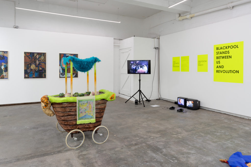 Photograph of a gallery, including a sculpture on wheels, a screen showing a film and artworks hung on the walls.