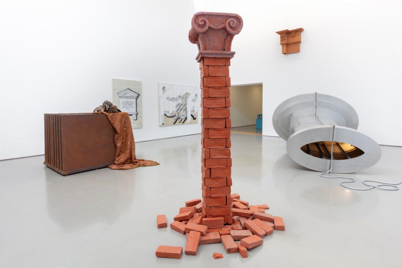 A series of sculptures in a white gallery. In the centre, a column made of brings that disintegrates into a pile of bricks at its base. in the background several large geometric shapes on their sides.