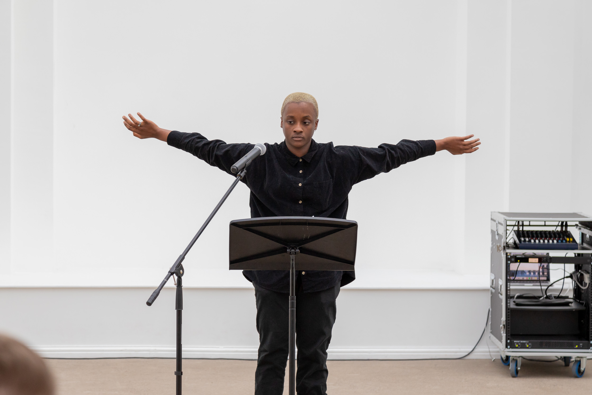 A black artist or performer stands at a microphone and music stand, their arms are flung wide. The background is white.