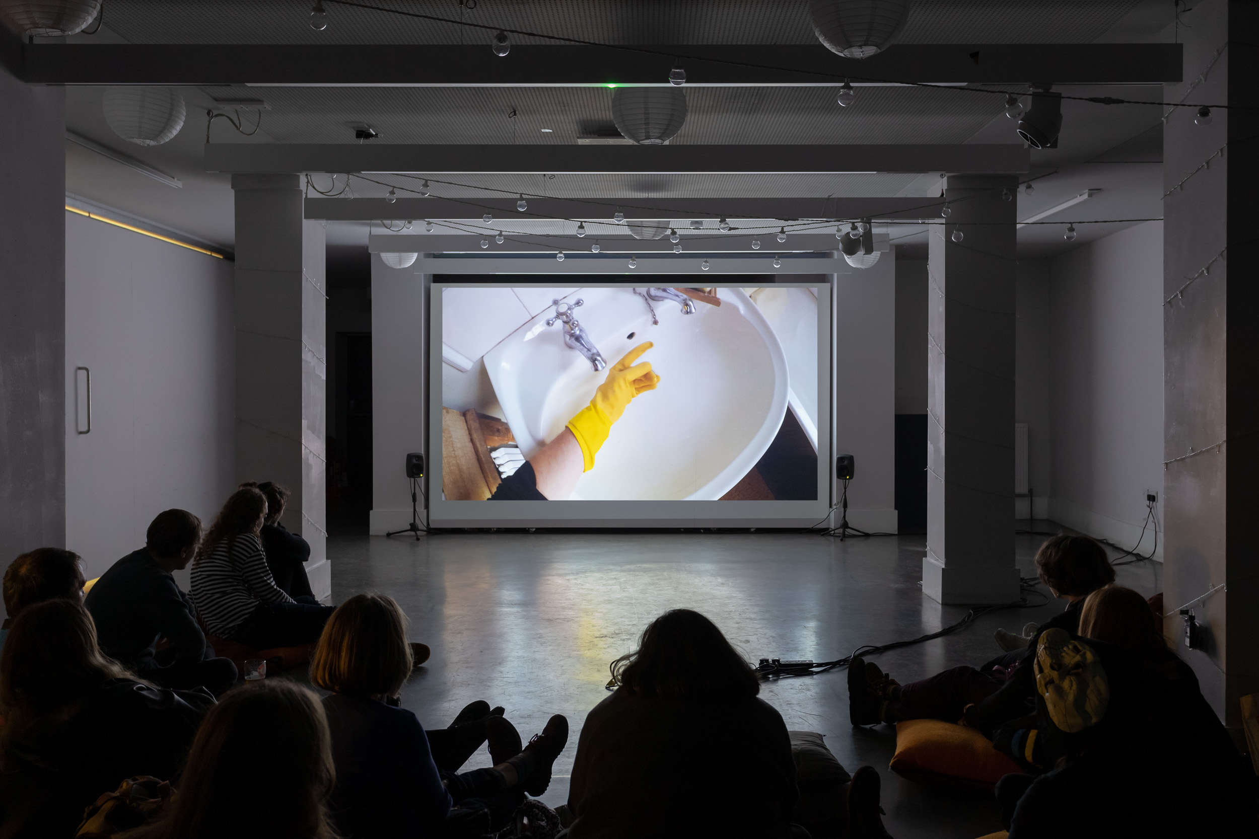 A film screened in a gallery, with people gathered to watch. On screen a hand in a yellow rubber glove points towards a white porcelain sink.