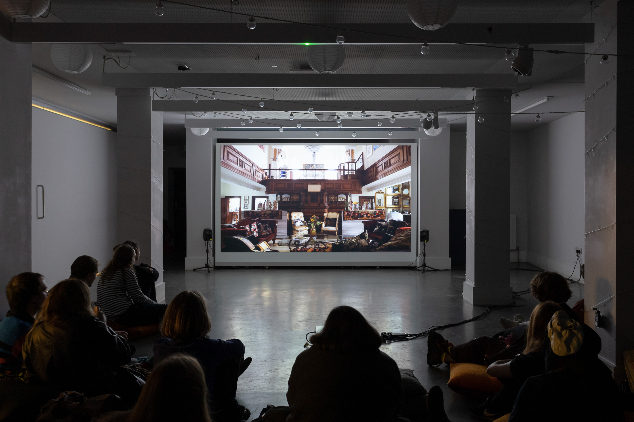 A film being screened in a gallery, with people gathered around to watch. On screen is a room filled with wooden furniture.