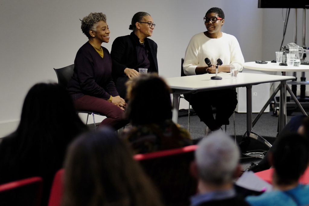 Three black women smile during a panel discussion. They are seated with a microphone and cups of water at a white table. The audience are visible in the foreground.