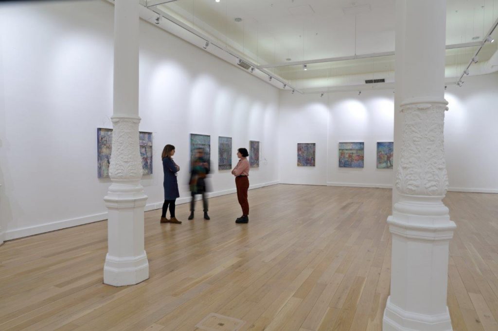 A white wall gallery space with a lightwood floor and white pillars. Works are hung on the walls, and several people are gathered in the space.