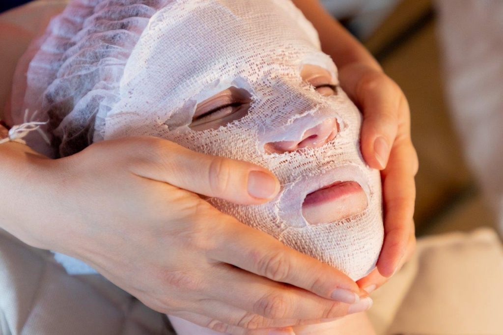 A person's face wrapped in a face mask and bandages, with their hair covered by a cap. Two hands hold either side of their face to massage it.