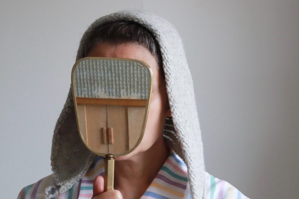 A woman holds a vanity mirror to obscure her face, The reflection in the mirror shows the doors of a dolls house. The woman has a grey knitted head piece on which falls to her shoulders. She is wearing a stripey pyjama top.