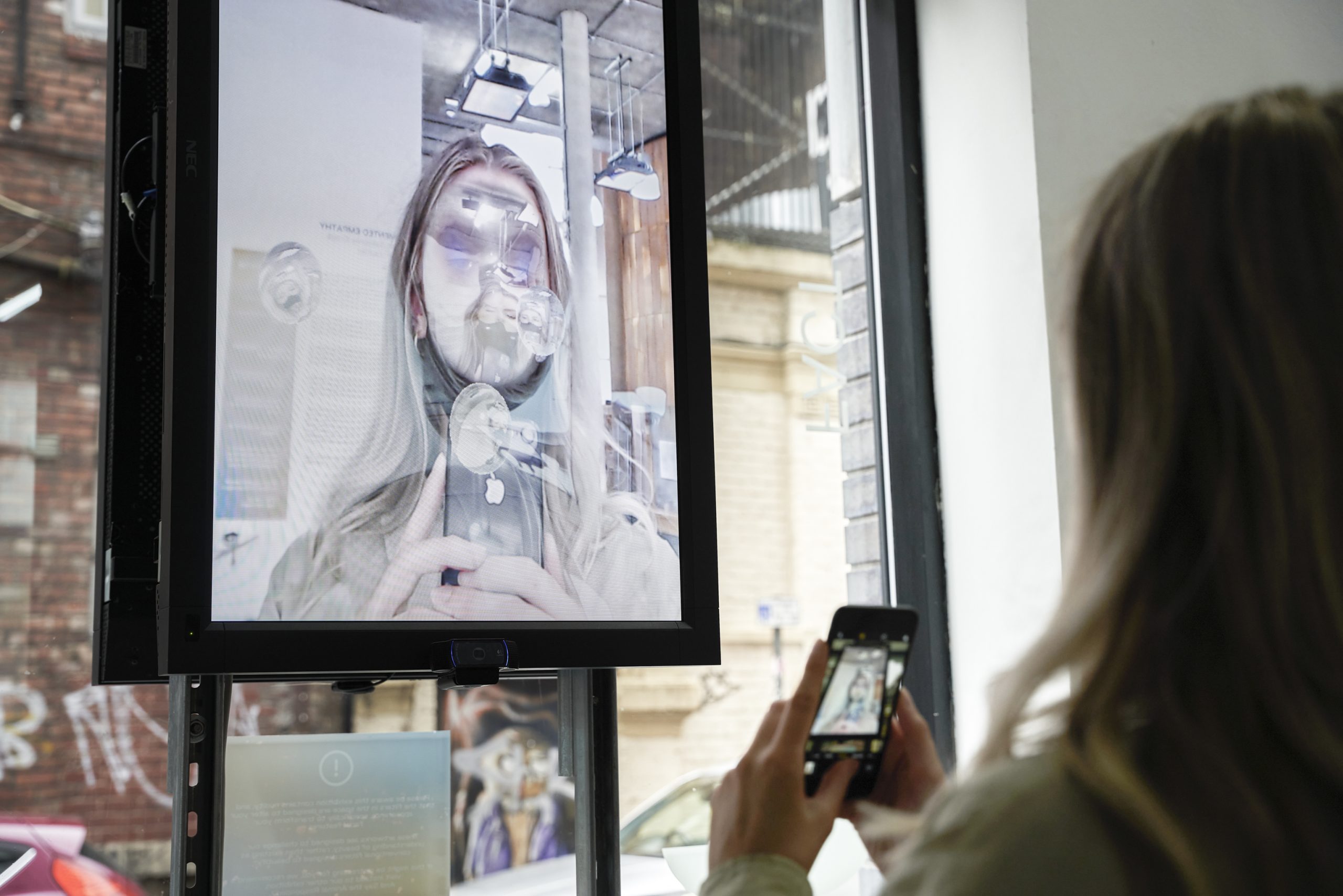 Someone takes a photograph of a framed image of themself, their face distorted with silver bubbles before it.