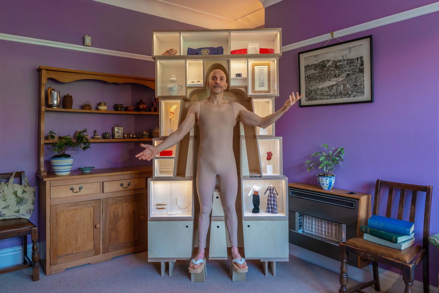 A person in a mauve bodysuit stands in a living room with purple walls. Behind them are shelves with objects, to the side is a wooden dresser with shelves, a framed print, an old style gas fire topped with a potted plant.