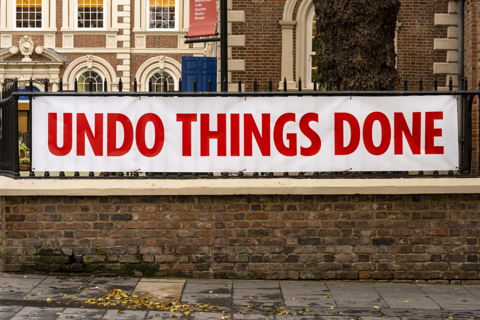 White banner with red text reading 'UNDO THINGS DONE' installed on black railings over a red-brick wall.