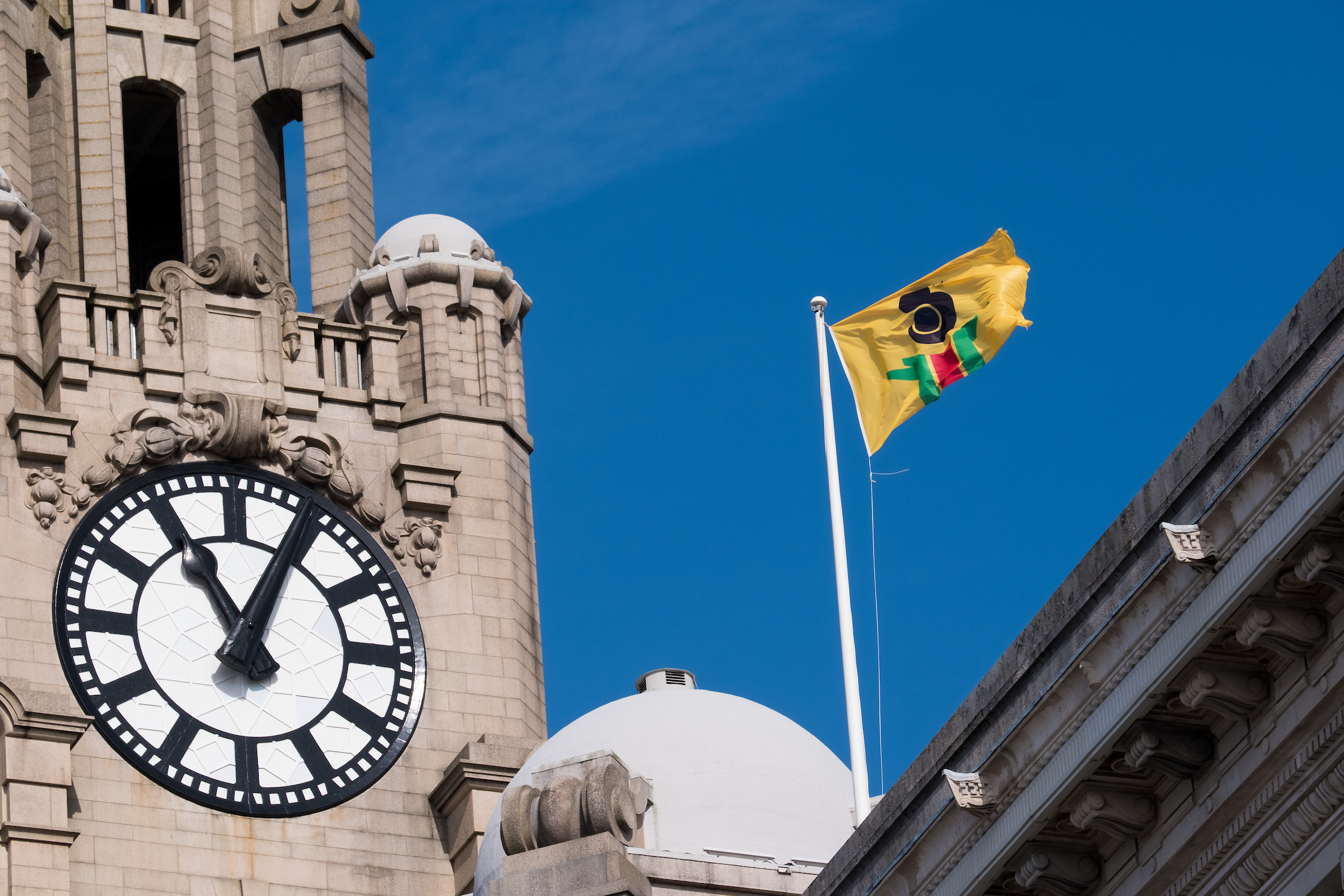 The yellow flag with green, red and black blocks flies against a bright blue sky next to a clock tower of the Royal Liver Building. The clock face shows the time as five past eleven. 