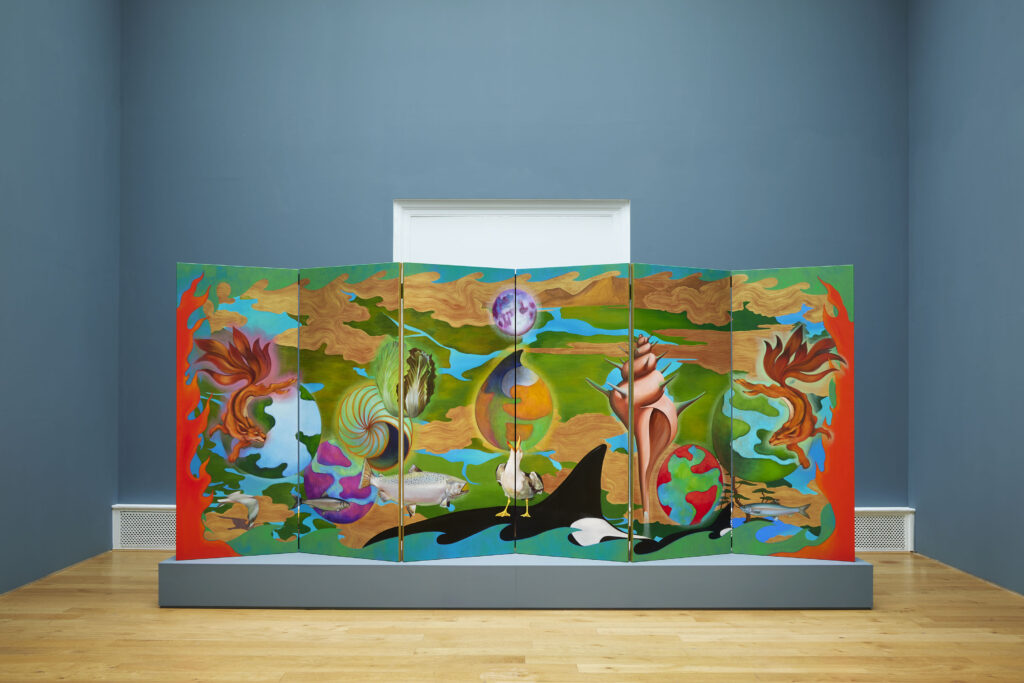 View of a large folding screen presented on a plinth in a gallery. The wooden screen is painted with images of animals, vegetables shells, waves, patches of land and planets. The colours are green, blue, with warm tones of red, orange and peach. The surrounding gallery walls are a dusky blue.