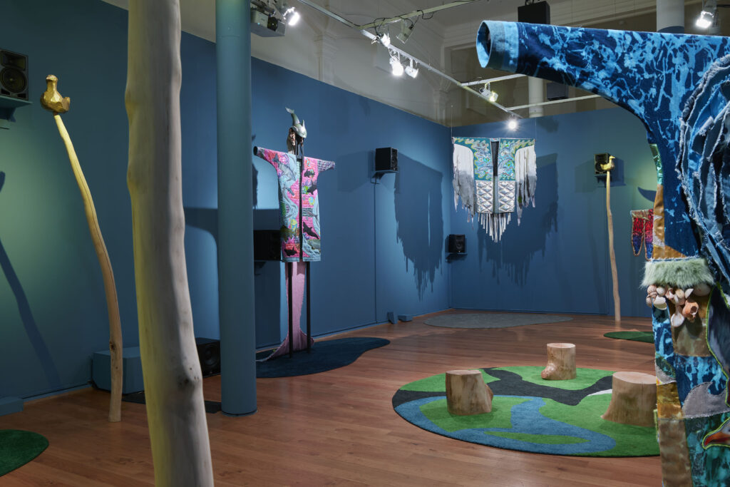 An installation view in a gallery. Several stitched garments are hung around the room, their arms outstretched. In the centre of the room there are tree stump seats on a green and blue rug. Around the blue walls are black speakers to play a sound piece.